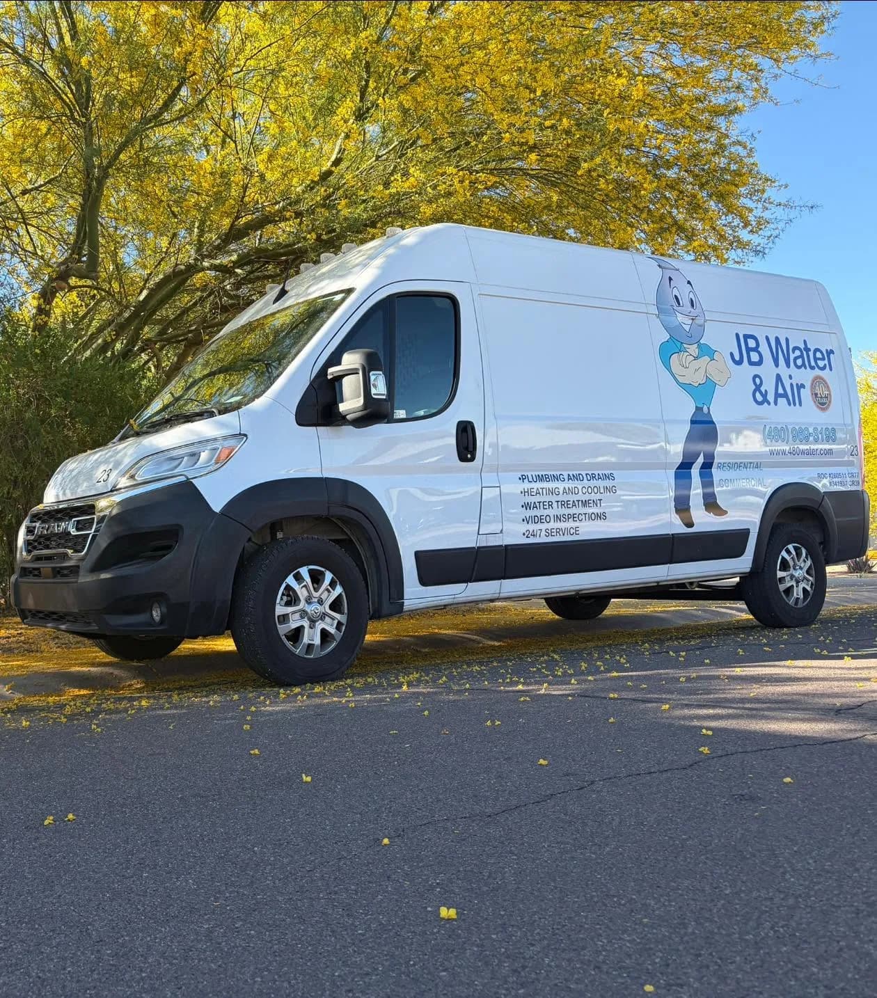 JB Water & Air's white Ram ProMaster service van, wrapped with the droplet mascot and the company phone number, parked under a flowering palo verde tree